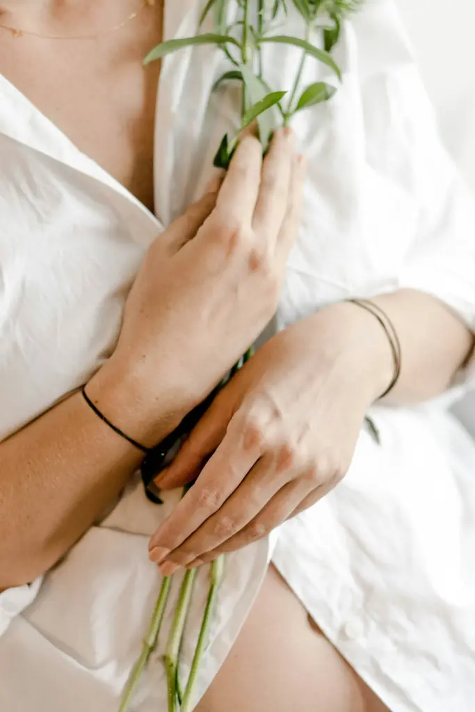 Eine schwangere Frau in weißem Hemd von schräg oben fotografiert, sie legt beide Hände über ihren Oberbauch und hält eine Pflanze zwischen Bauch und Hände.